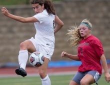 A woman in white in the foreground in midair about to kick a soccer ball while a defensive player readies herself. 